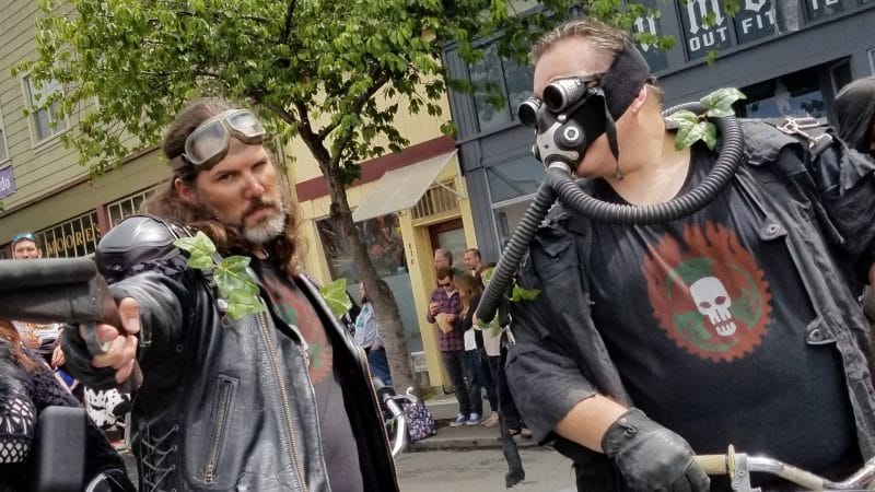 Two men dressed in leather and gas masks on a motorcycle.
