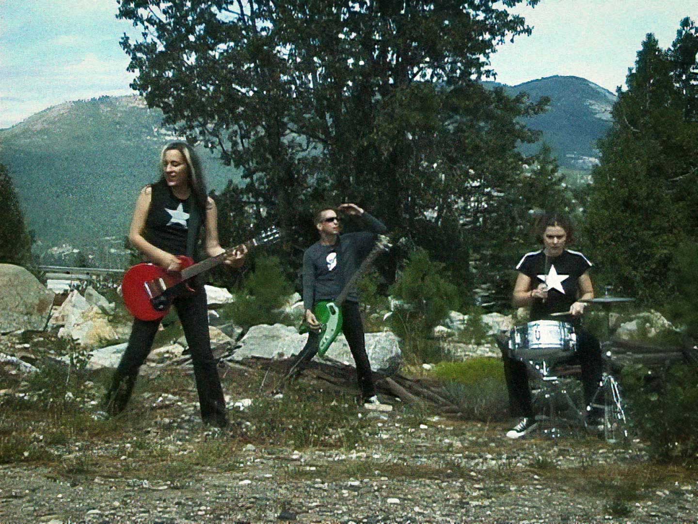 A group of people playing guitars and drums in front of a mountain during a Groovie Ghoulies Music Video.