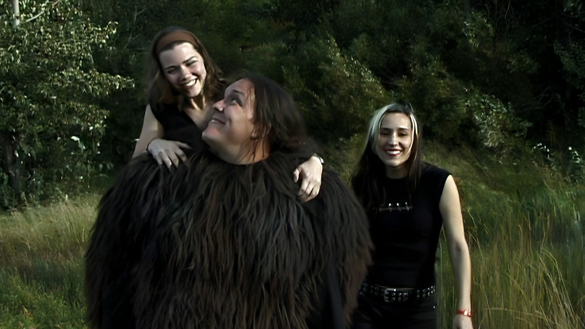 Two women standing in a field with a large furry creature in a Groovie Ghoulies music video.