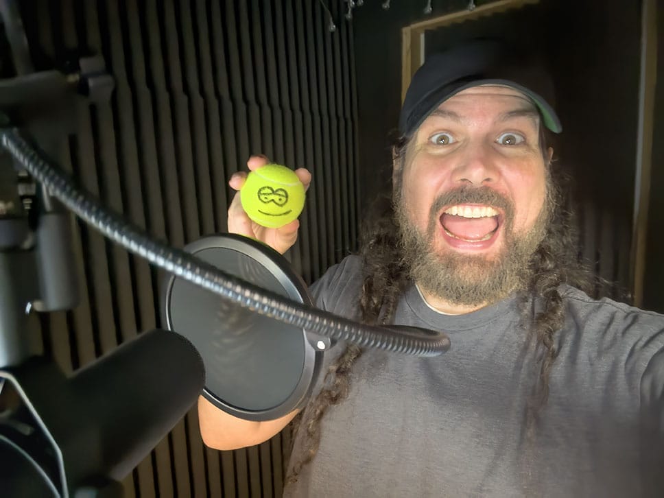 Man with long hair and a beard smiling broadly in a soundproof booth, holding a green stress ball with a smiley face, near a microphone and pop filter.