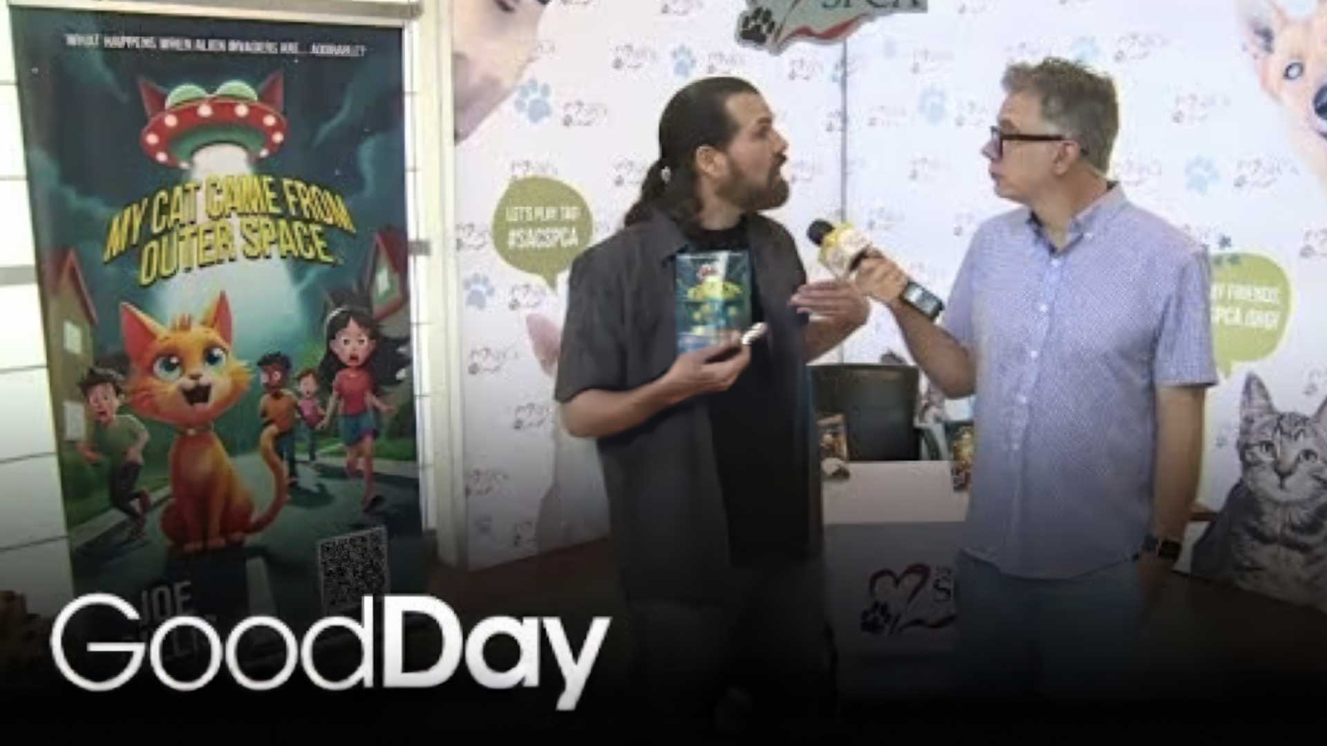 Joe Gillis and Cody Stark talk during a “Good Day Cody's Caravan” interview, Joe holding his "My Cat Came From Outer Space" book, and Cody holding the microphone towards Joe. A “My Cat Came From Outer Space” poster and a cat-themed backdrop are visible behind them.